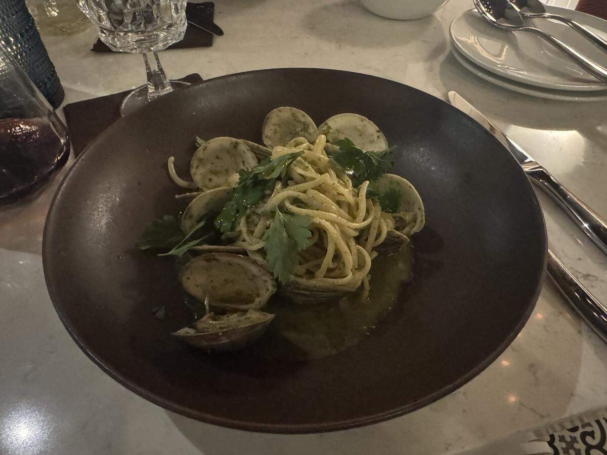 A dark brown ceramic bowl filled with a pasta dish, positioned on a marble table. Long strands of spaghetti or linguine are tossed in a light green herb sauce, mixed with several open clams in their shells. The dish is garnished with fresh flat-leaf parsley leaves.
