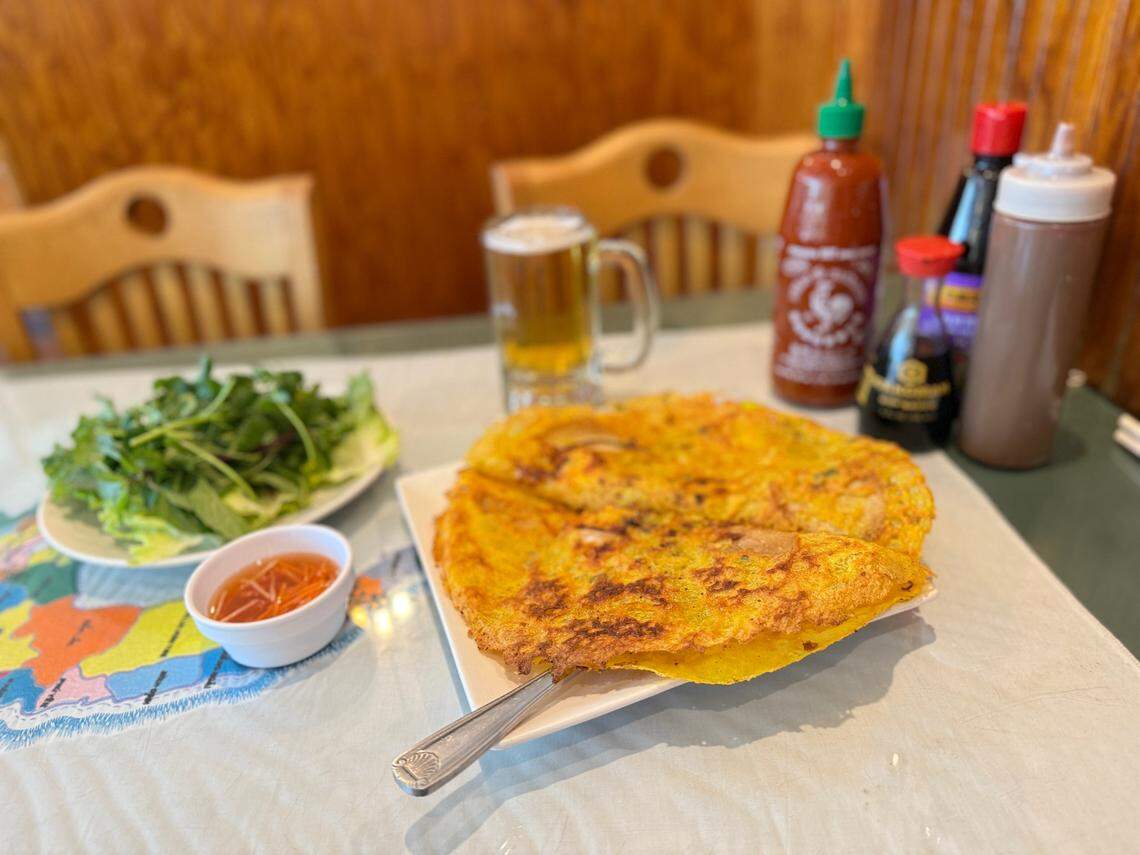 A high-angle, close-up shot of a Vietnamese crepe (bánh xèo), a large, golden-yellow, crispy crepe folded in half on a white plate. The meal is set on a table with a map-themed tablecloth, accompanied by a small bowl of dipping sauce, a plate of fresh herbs and lettuce, a mug of beer, and various condiment bottles like Sriracha and soy sauce in the background.