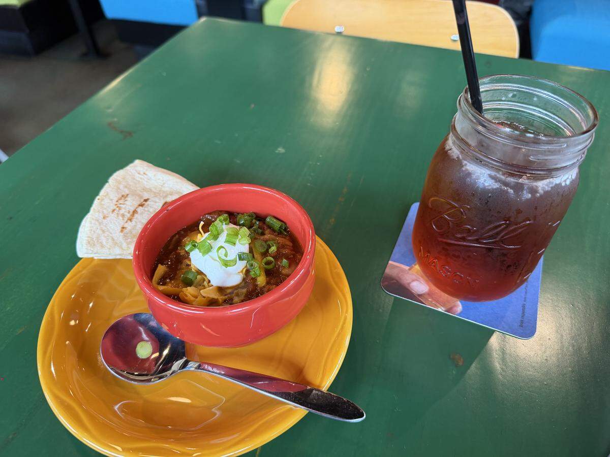 A small red bowl of chili topped with shredded cheese, sour cream, and green onions, served on an orange plate with a side of flour tortillas. Next to it sits a large mason jar filled with iced tea and a black straw, resting on a blue coaster.