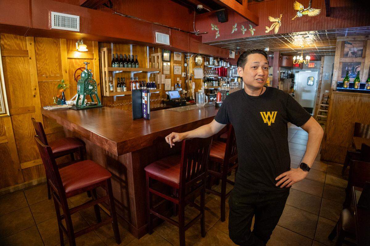 A restaurateur in a black t-shirt with a yellow logo stands leaning against a dark wood bar in a Thai restaurant. The background shows wine bottles on shelves, a vintage ice shaver, and gold bird decorations on the wall.