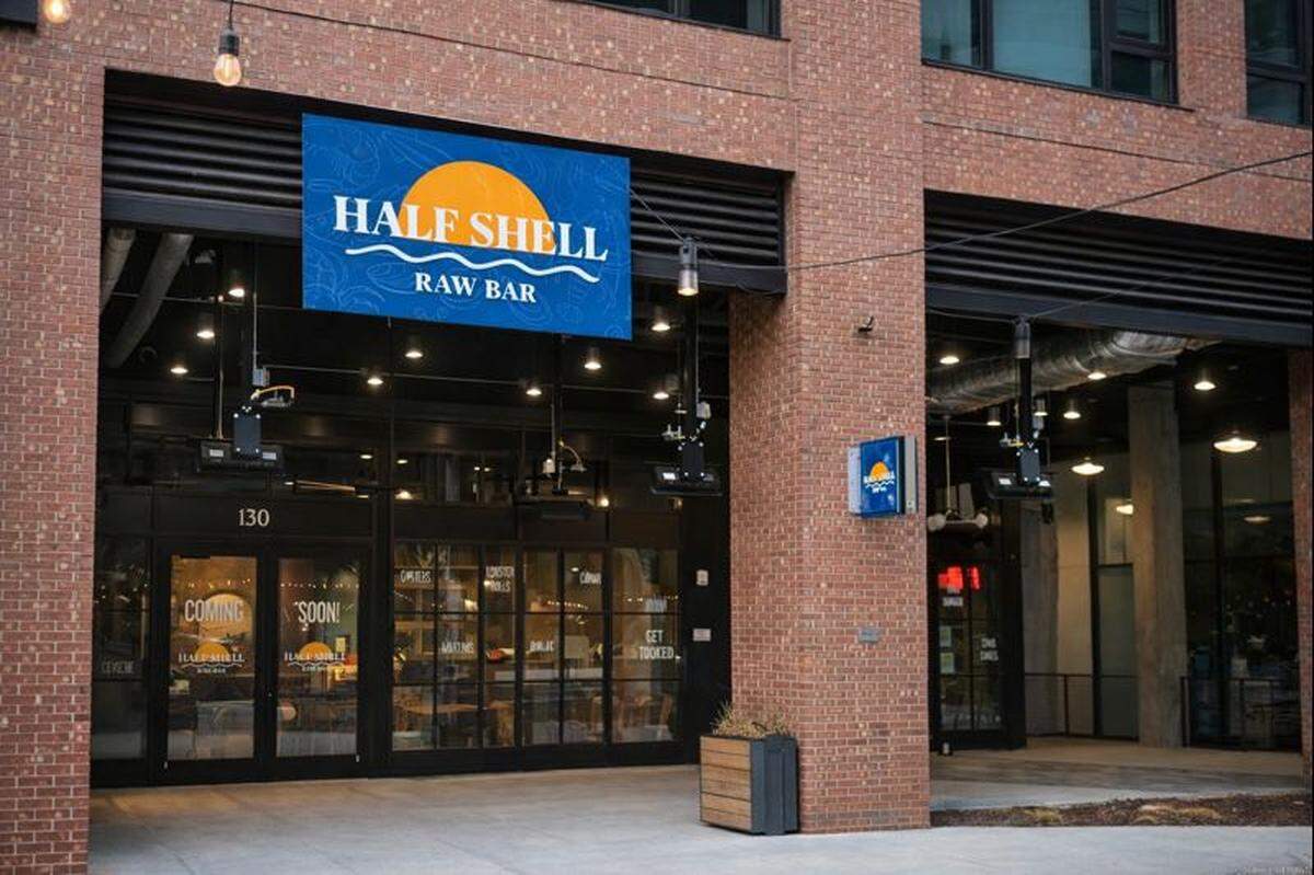 The exterior of “Half Shell Raw Bar” features a modern brick facade with large glass windows and a blue-and-orange sun logo. The entrance is well-lit with hanging industrial lights and a “Coming Soon” sign visible on the front door.