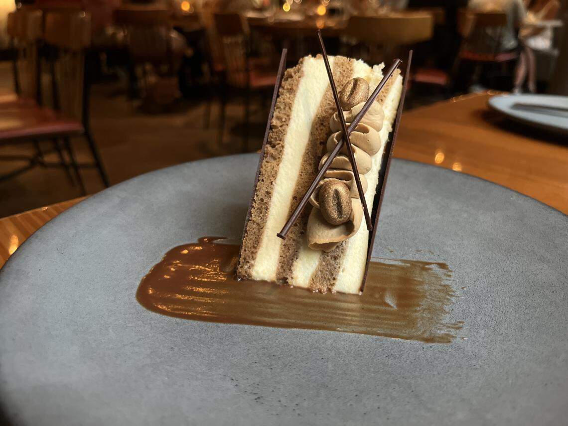 A dessert plate holds a rectangular slice of layered cake. The cake has alternating layers of light and dark sponge cake and a creamy filling. It is garnished with several chocolate sticks and dollops of a tan-colored cream. The slice of cake stands upright on a gray plate, which is swirled with a brown caramel-like sauce. The background is a blurred view of a restaurant.