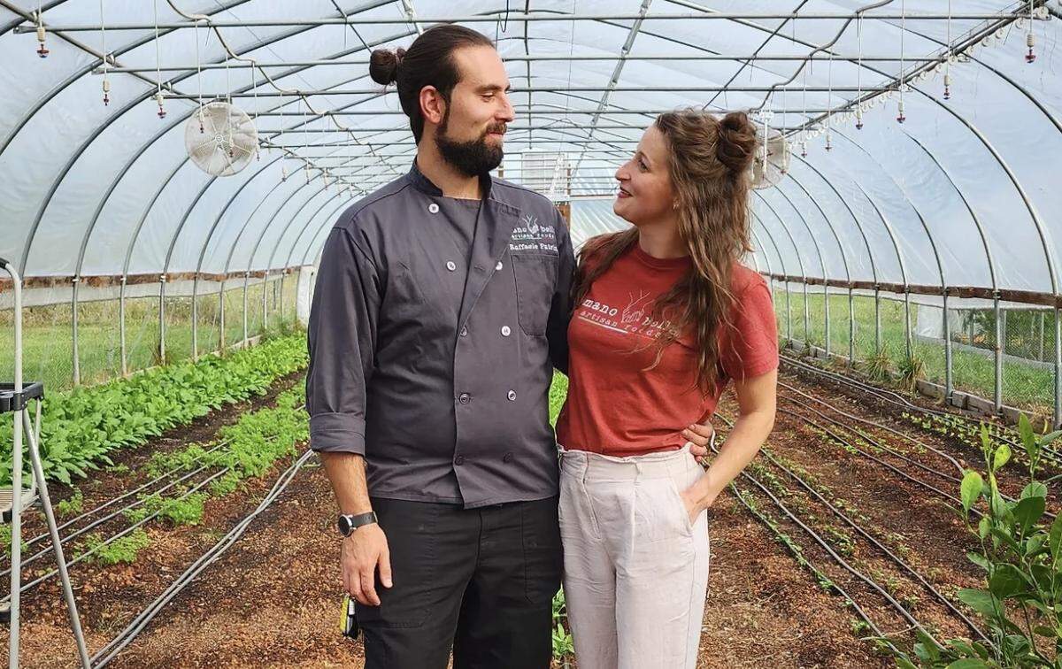 A medium shot of a smiling couple looking affectionately at each other inside a large hoop house greenhouse. The person on the left wears a dark gray chef’s coat, and the one on the right wears an orange logo t-shirt and light linen pants. They stand along a central dirt path, flanked by neat rows of green crops with drip irrigation lines under the curved metal frame of the structure.