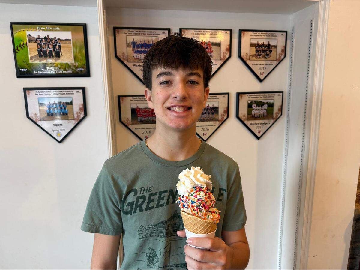 A young person holding a tall waffle cone filled with ice cream and completely covered in multi-colored rainbow sprinkles, finished with a swirl of whipped cream. The teenager is standing against a white wall decorated with several diamond-shaped plaques honoring youth athletes.