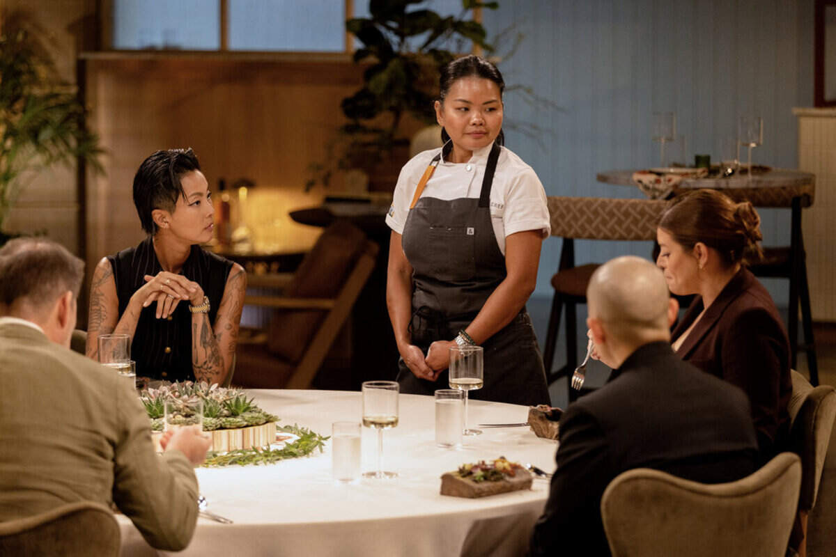An eye-level shot from a television show set showing a female chef standing and talking to a panel of judges seated around a large white dinner table. In the center, the chef stands in a white short-sleeved chef’s coat and a charcoal gray apron, looking intently off to her right. Seated around the foreground table are several judges, including Top Chef host Kristen Kish on the left, who has slicked-back short black hair, visible arm tattoos, and wears a sleeveless pinstriped top while leaning forward with her hands clasped. To the right, Gail Simmons is partially visible in profile, wearing a dark blazer, while a bald male judge sits next to her with his back mostly to the camera.