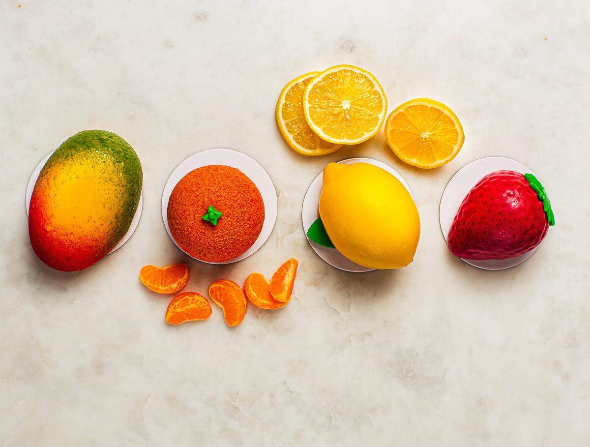 A top-down view of four gourmet desserts shaped like realistic fruit—a mango, an orange, a lemon, and a strawberry—resting on a white marble surface. Real slices of citrus fruit are scattered around the center desserts.