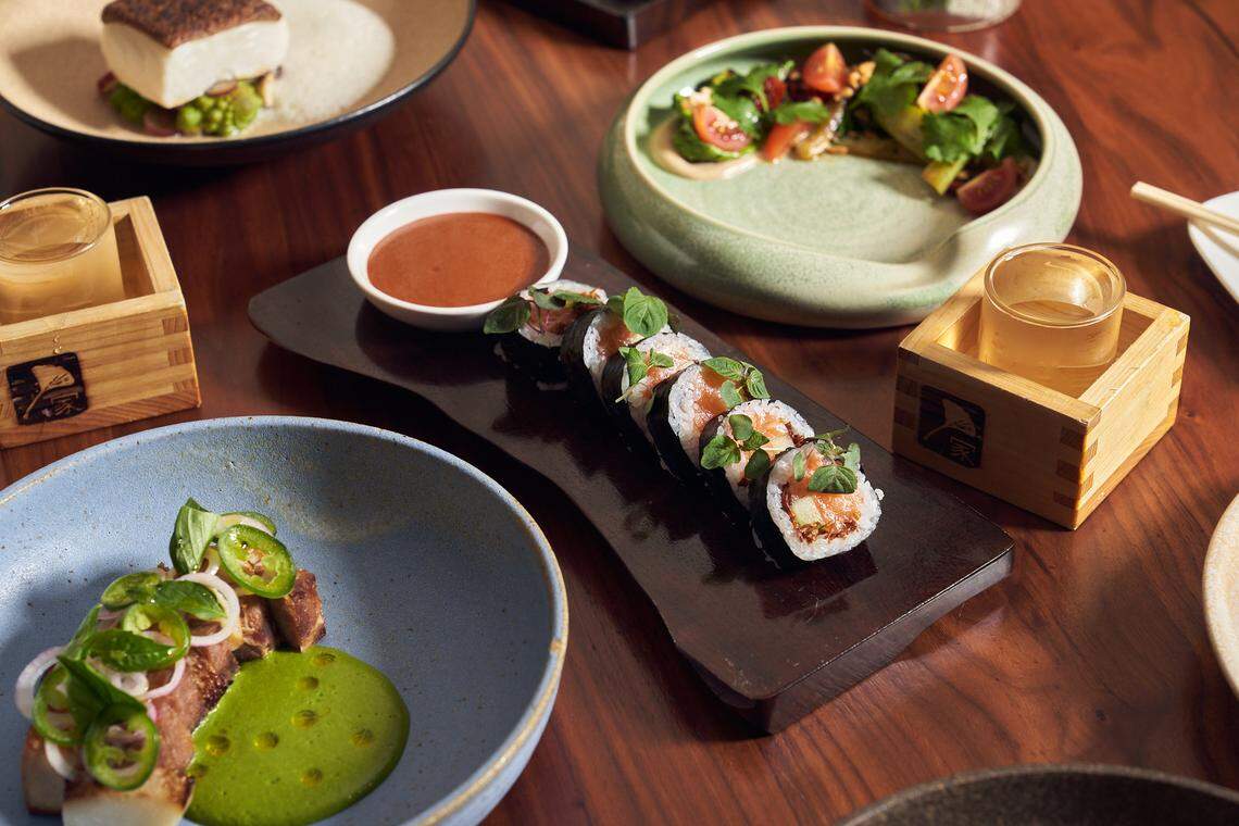 A high-angle view of a wooden table spread with various Japanese-inspired dishes. In the center is a dark rectangular plate with a row of sushi topped with fresh green herbs. Surrounding it are small bowls containing a vibrant green pea purée with pork belly, a salad with cherry tomatoes, and a cooked white fish fillet. Two small glasses of amber liquid are served in traditional wooden masu boxes.