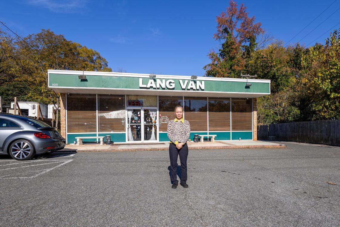 A wide, straight-on shot of a restaurant owner standing in the middle of a parking lot in front of the “Lang Van” restaurant on a sunny day. The person is in the center of the frame, wearing a zebra-print cardigan, and looking at the camera. The one-story restaurant building, featuring a large green sign with white letters, fills the background under a clear blue sky.