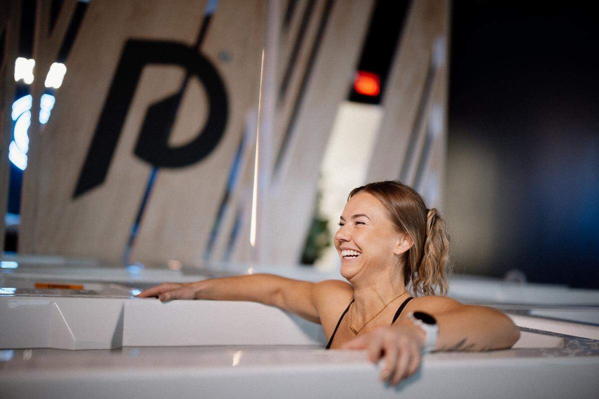 A medium shot of a person with blonde hair tied back in a ponytail, laughing and smiling while submerged in a white cold plunge tub with hrms resting on the edges of the tub. The background is slightly blurred, showing a modern interior with a large black “P” logo on a light wooden wall.