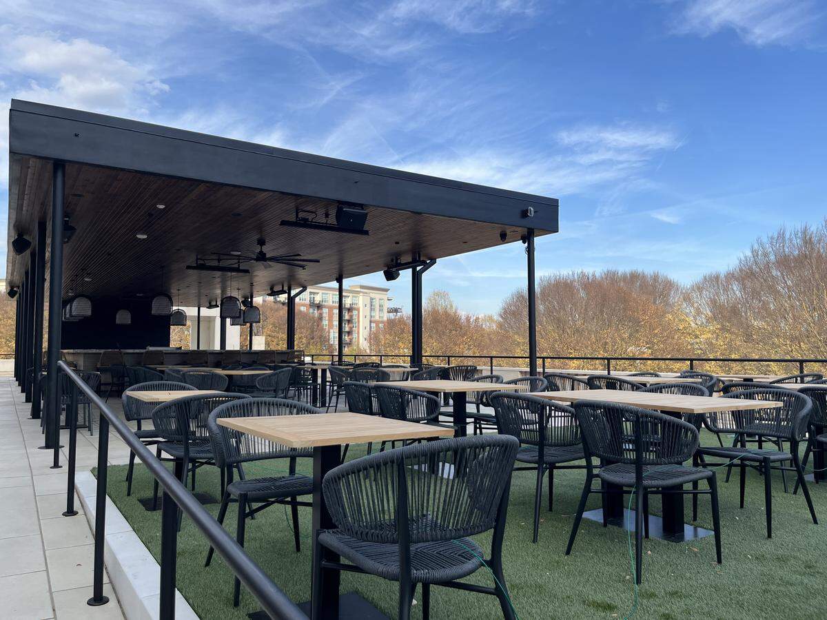 A wide, eye-level shot of a spacious outdoor dining terrace covered in artificial green turf. The patio is furnished with many light wood-topped tables and black woven rope chairs. A black covered structure with a wood plank ceiling is on the left, and the terrace overlooks trees with yellow autumn leaves under a bright blue sky.