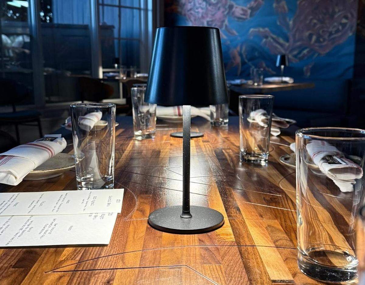 A long wooden dining table set for a group, featuring water glasses and white napkins with red stripes. A sleek, black cordless table lamp stands in the center of the table. In the background, a blue mural is visible on the wall.