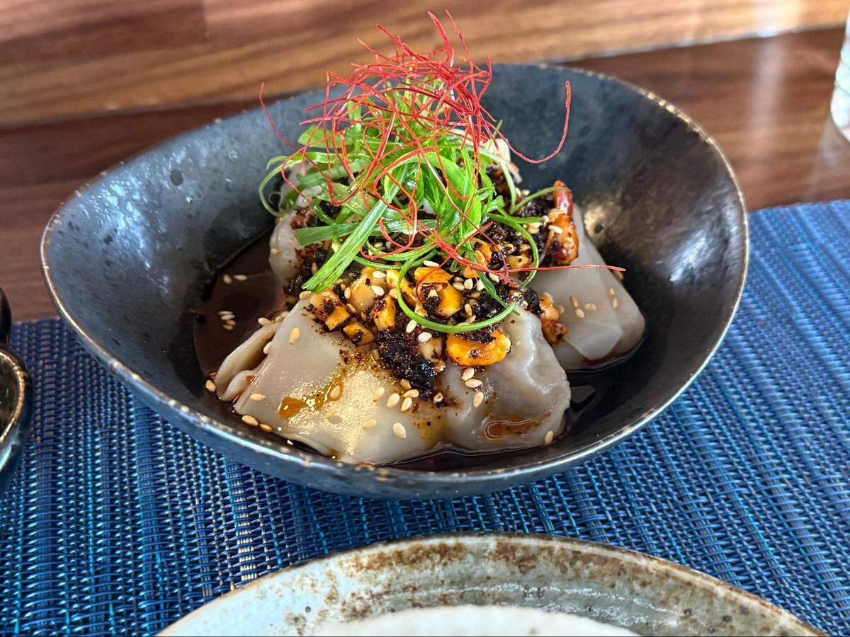 Three silky, translucent steamed dumplings in a shallow black bowl, bathed in a dark soy and red chili oil sauce. The dumplings are garnished with toasted sesame seeds, chopped peanuts, sliced green scallions, and thin red chili threads.