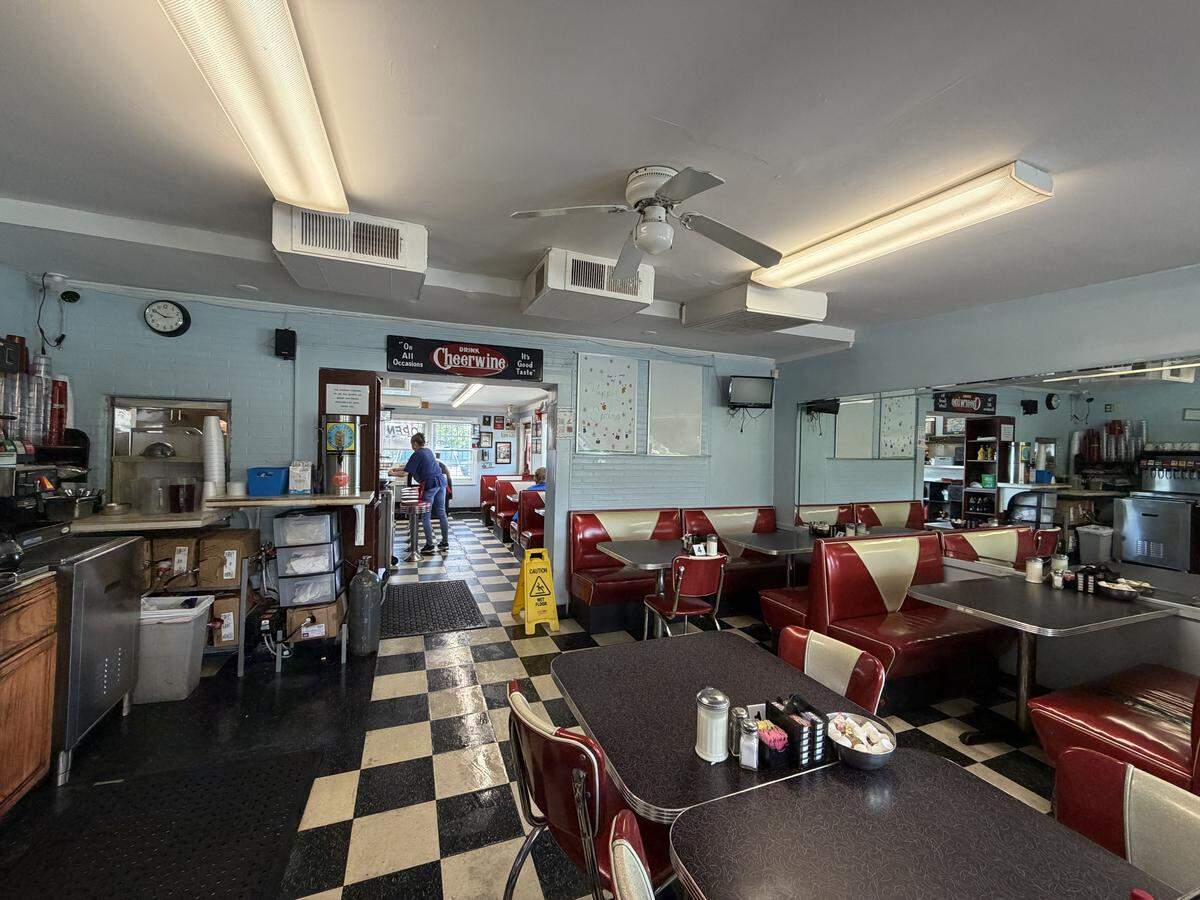 A wide interior view of a classic American diner with light blue walls and a black-and-white checkered floor. The room is filled with red-and-white vinyl booths and speckled grey tables. To the left, a small service area contains coffee equipment and storage. A doorway in the center leads to another seating area where a person is visible near the counter. A vintage “Drink Cheerwine” sign hangs above the passage, and a white ceiling fan is centered between long fluorescent light fixtures.