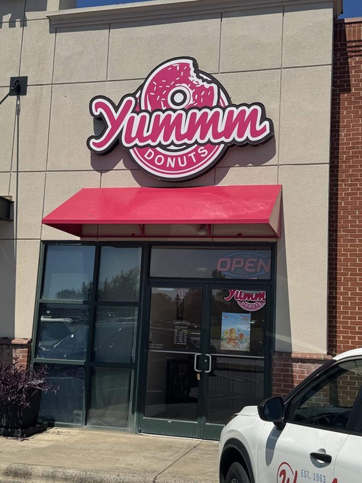 A head-on view of the storefront for YUMMM Donuts. A large sign with a half-eaten doughnut and the store’s name in pink and white letters is mounted on the light gray building above a red awning. Below the awning are the store’s glass doors and windows, with a white truck parked to the right.