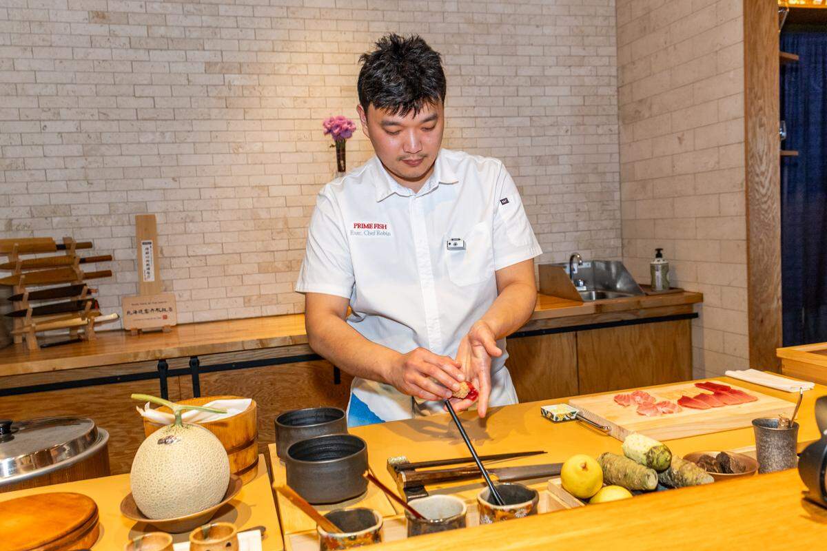 A chef stands behind a wooden sushi counter, looking down while carefully shaping a piece of nigiri. The counter is organized with various ingredients, including a whole cantaloupe, several small bowls, and a wooden board with slices of raw tuna.