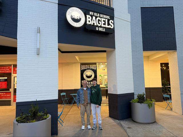 An outdoor, eye-level shot shows two people standing in front of a shop named NY Old School Bagels & Deli. On the left, a person with gray hair wears a denim jacket over a brown shirt, tan cargo pants, and white sneakers. To the right, a taller and younger person with dark hair wears a green hooded sweatshirt, light gray sweatpants, and gray sneakers. They are both smiling at the camera.