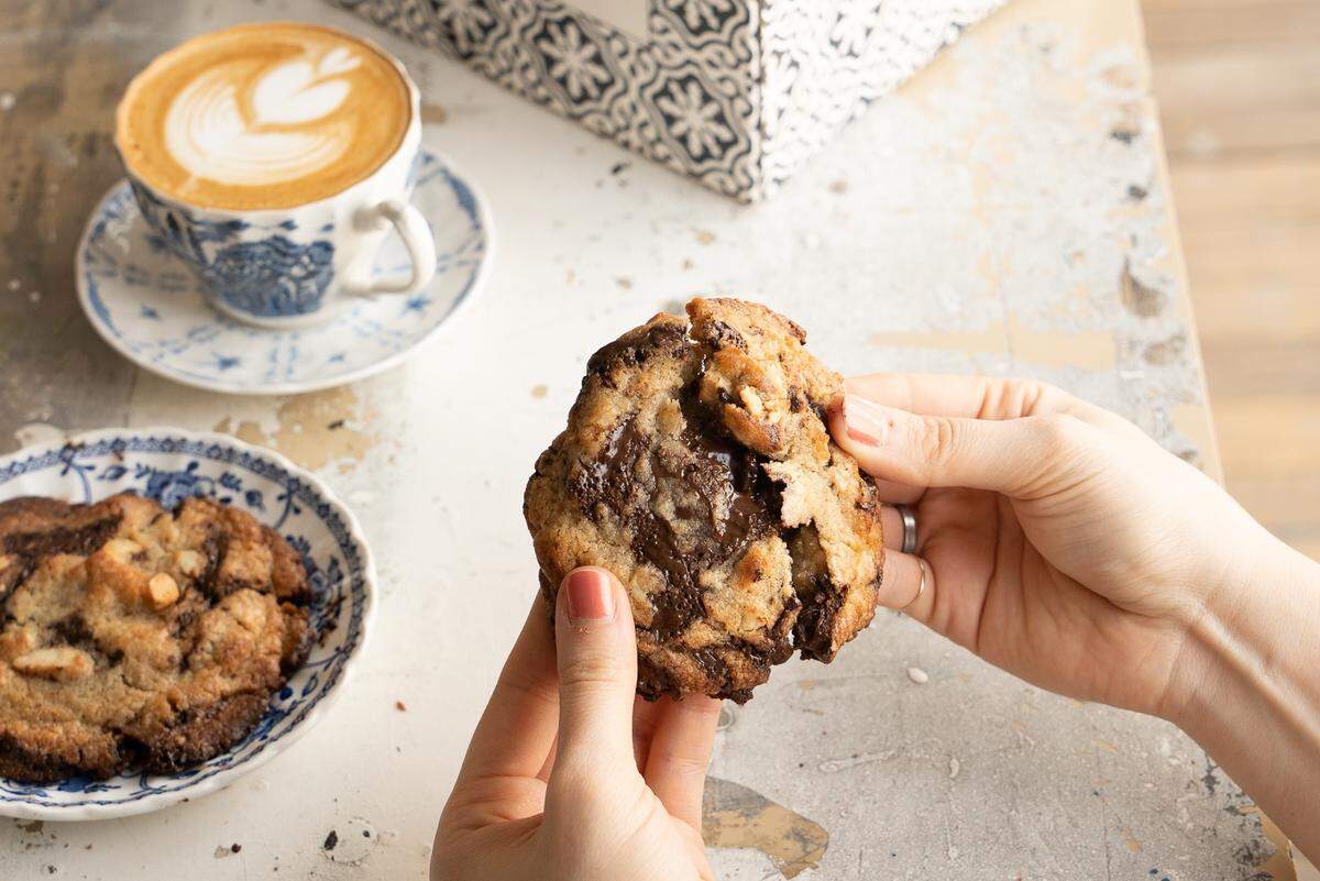 A person’s hands break open a thick, gooey chocolate chip cookie filled with melted chocolate and nuts over a rustic white tabletop. In the background, a heart-patterned latte in a blue-and-white teacup sits beside another cookie on a matching saucer.