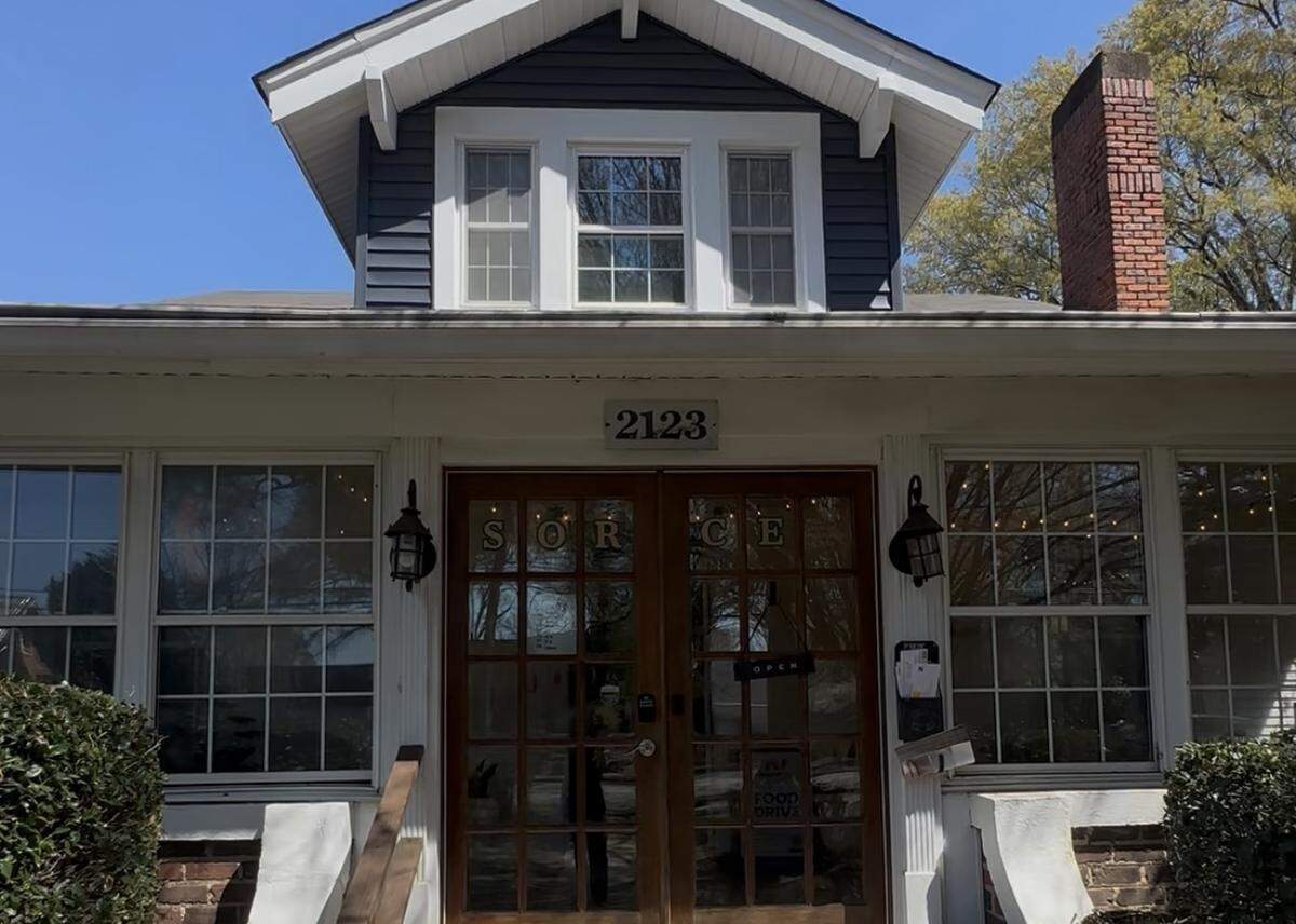The front exterior of a charming, two-story cottage converted into a shop. It features a dark blue gabled dormer with three windows and a red brick chimney. The number “2123” is displayed above the front French doors, which have the word “SORCE” applied in gold lettering across the glass panes.