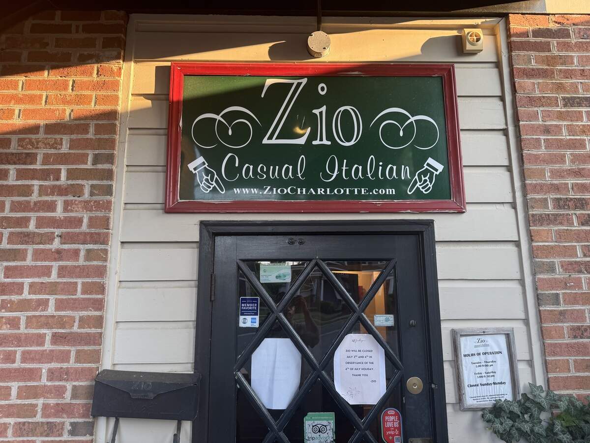 A close-up of the front entrance of a restaurant. A dark wooden door with diamond-patterned window panes features a white paper sign taped to it regarding holiday hours. Above the door is a dark green sign with a red frame that reads “Zio Casual Italian” with the website address below. Two illustrated hands point down toward the entrance. The building’s exterior is made of light-colored siding and red brick, illuminated by warm, late-afternoon sunlight.