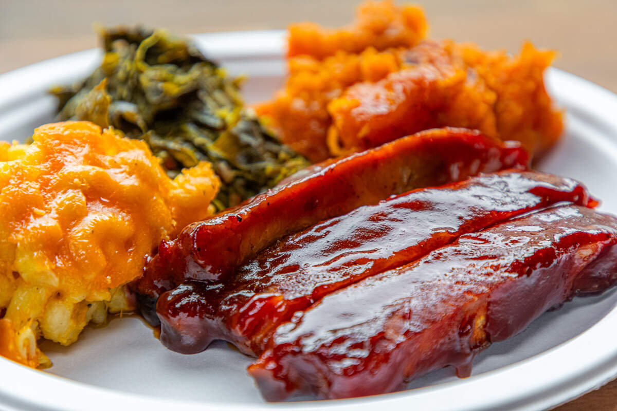 A close-up of a white plate holding three pork ribs heavily glazed in a glossy, dark red barbecue sauce. The ribs are served alongside a scoop of creamy baked macaroni and cheese, a portion of slow-cooked collard greens, and a side of mashed candied sweet potatoes.