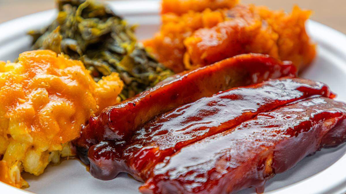 A close-up of a white plate holding three pork ribs heavily glazed in a glossy, dark red barbecue sauce. The ribs are served alongside a scoop of creamy baked macaroni and cheese, a portion of slow-cooked collard greens, and a side of mashed candied sweet potatoes.