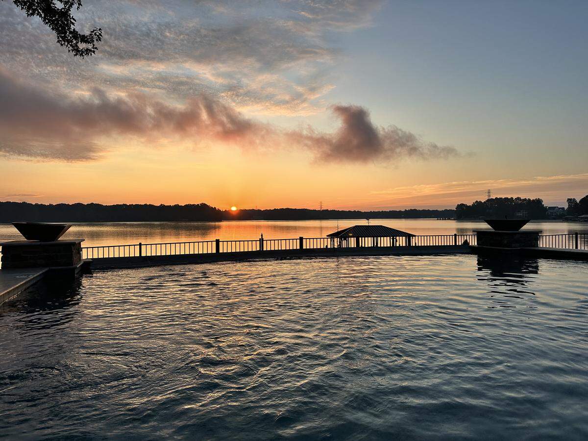 A peaceful sunrise over a lake is viewed from behind an infinity pool. The sun is a bright orange orb on the horizon, under a sky with pink and orange clouds. The pool’s water in the foreground reflects the light, and in the distance, a covered boat dock is visible on the calm lake.