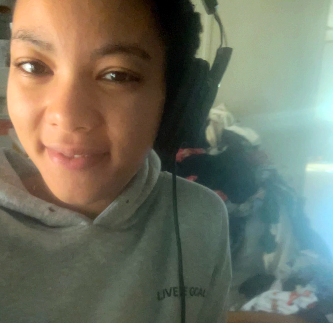 A close-up selfie of a smiling person wearing a gray hooded sweatshirt and over-the-ear headphones, looking directly at the camera. The background is out of focus, showing a pile of clothes.