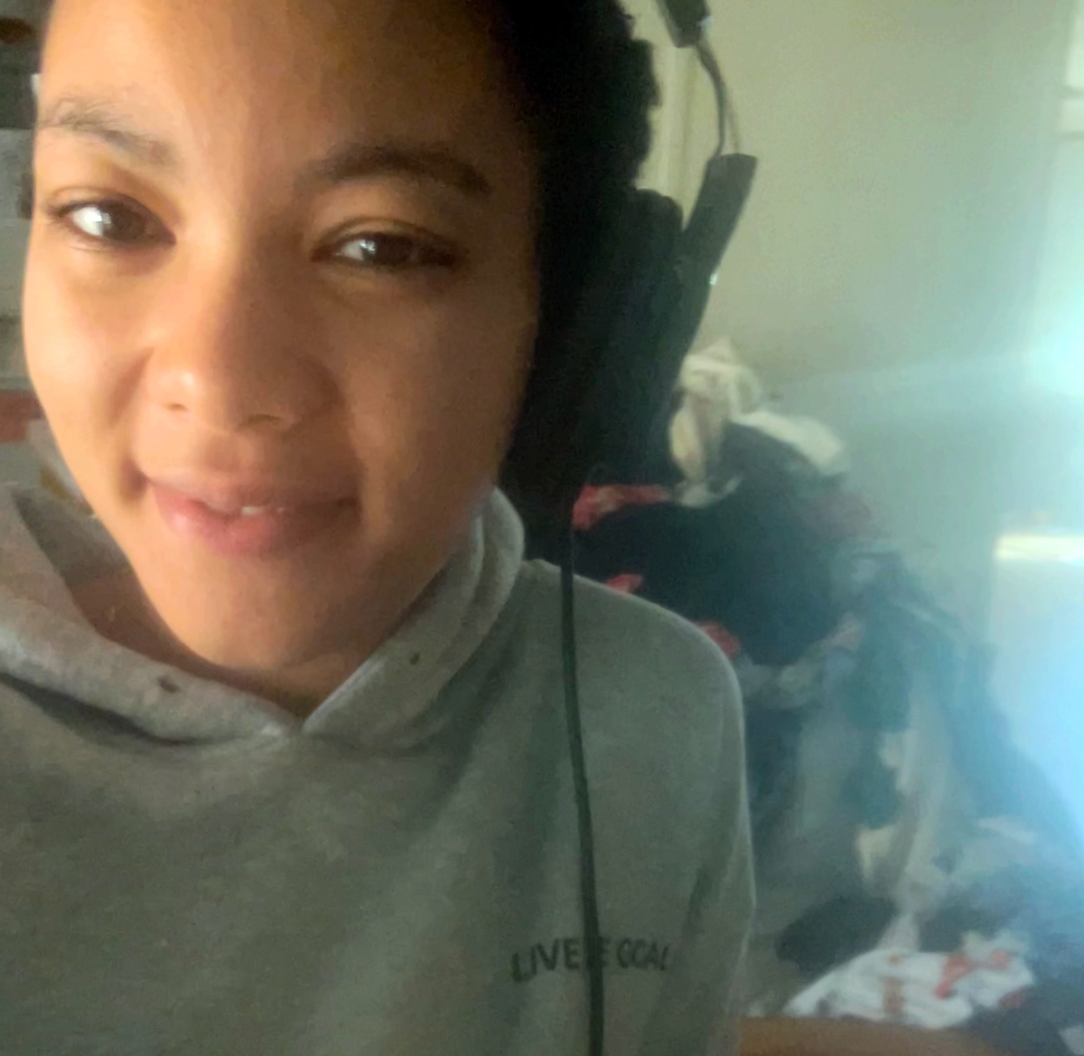 A close-up selfie of a smiling person wearing a gray hooded sweatshirt and over-the-ear headphones, looking directly at the camera. The background is out of focus, showing a pile of clothes.