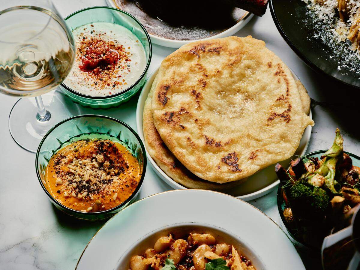 An overhead shot of several small plates of Mediterranean-inspired food spread across a white marble dining table. People’s hands are visible, eating with forks and knives. Dishes include shrimp in a red sauce, kofta skewers, falafel on leaves, and a salad. A glass of rosé wine and a glass of water are also on the table.