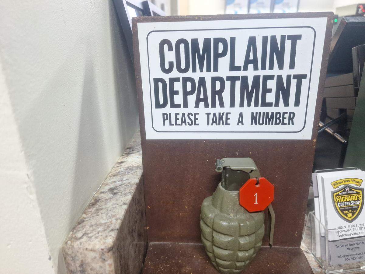 A playful sign in a business reads “COMPLAINT DEPARTMENT PLEASE TAKE A NUMBER” in bold black letters. Below the sign, a green replica hand grenade has a red tag with the number “1” hanging from its pin, serving as the “take a number” ticket. The grenade sits on a brown counter next to a stack of brochures for “Richard’s Coffee Shop” and “Welcome Home Veterans.”