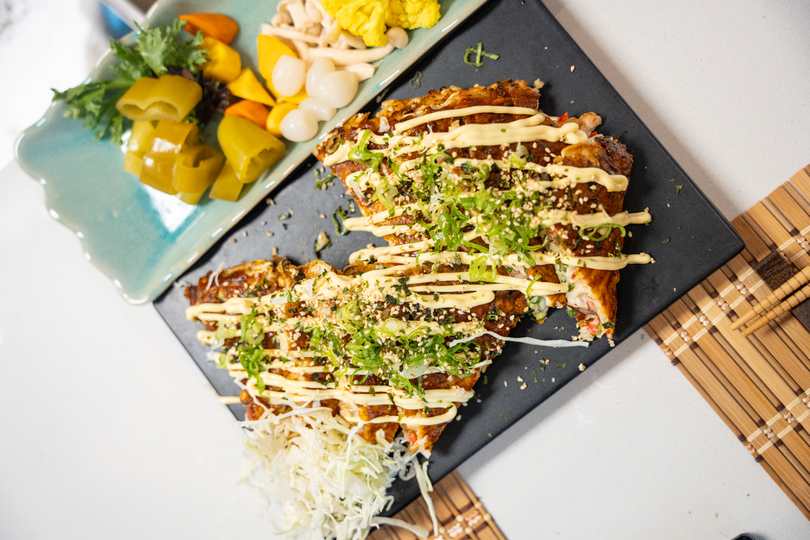A Japanese okonomiyaki pancake, drizzled with sauce and sprinkled with herbs, is served on a black slate platter. A side of shredded cabbage is on the platter, and a separate dish of assorted Japanese pickles is to the side.