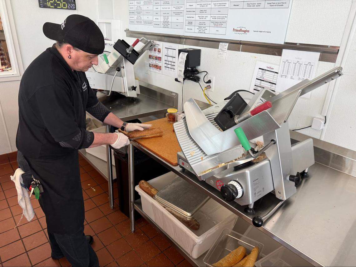 A sandwich shop worker wearing a black shirt, apron, and backwards baseball cap is standing behind a stainless steel counter, using a knife to slice a long sesame seed hoagie roll. Two large industrial meat slicers are visible on the counter.