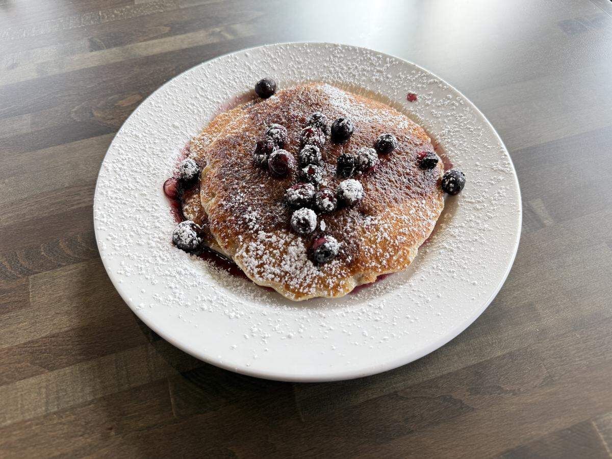 A white plate holding two golden-brown pancakes topped with a blueberry compote and dusted generously with powdered sugar, sitting on a dark wooden table.