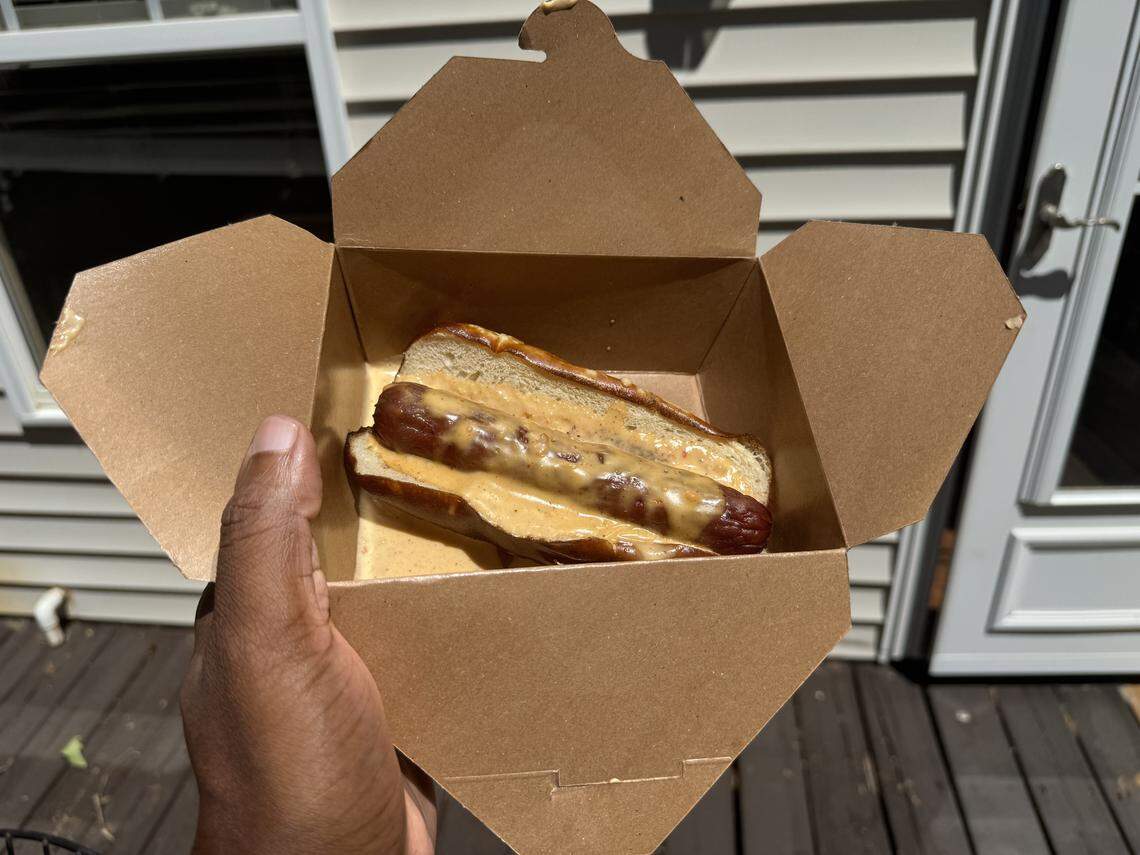 A hand holding an open brown cardboard takeout box containing a large grilled hot dog in a toasted bun, completely drenched in a thick, golden-orange cheese sauce.