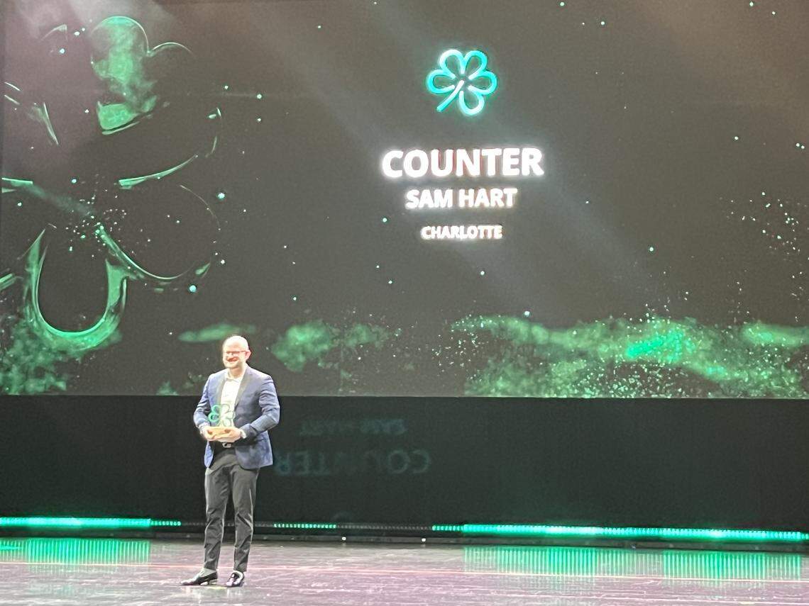 A wide shot of Sam Hart of “Counter” in Charlotte, standing alone on a large stage and smiling as they hold their Michelin Green. Star award. A large screen behind him displays their name, restaurant, and the Green Star logo.