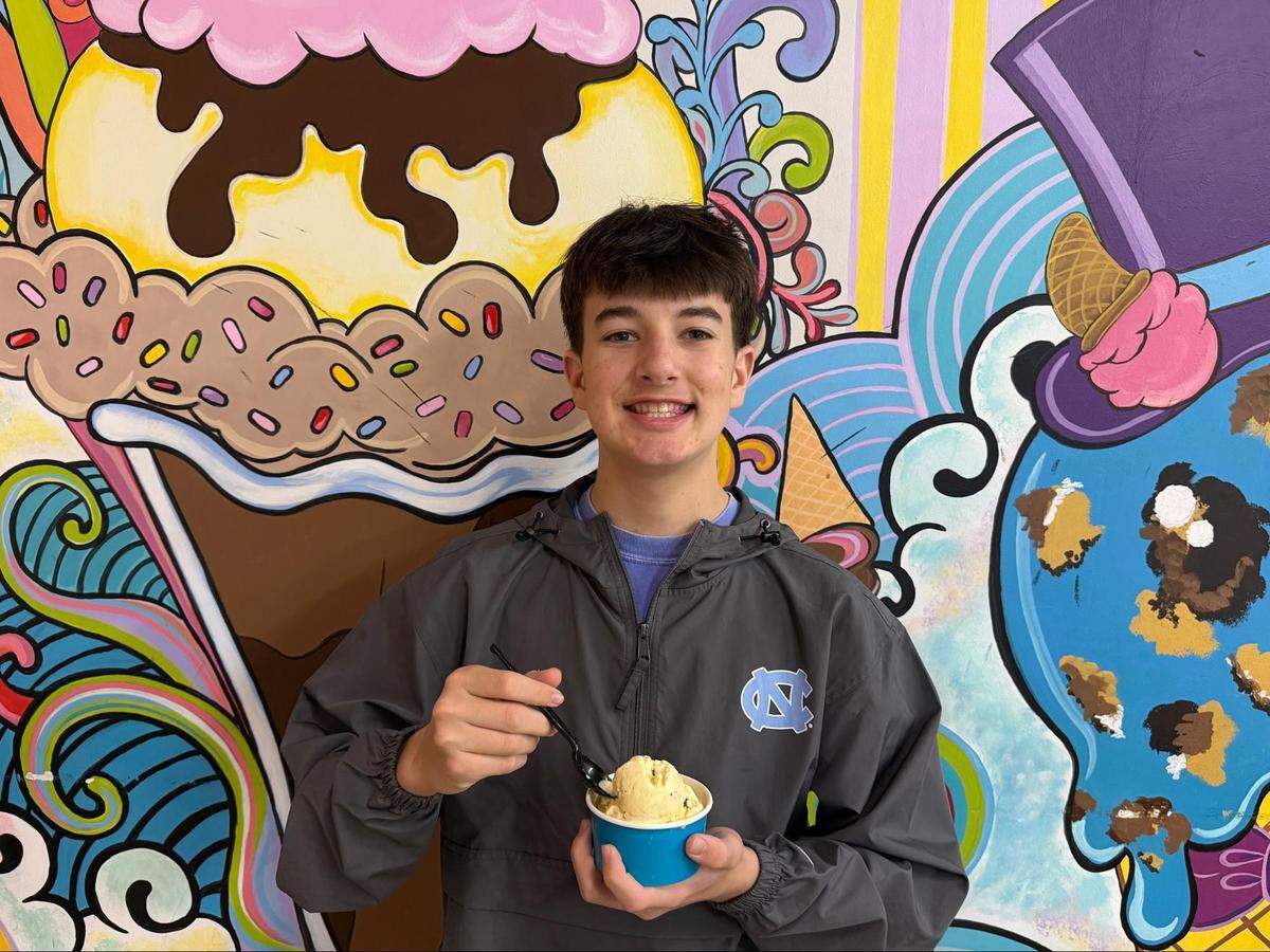 A young man in a grey UNC windbreaker smiles while holding a small blue cup of pale yellow ice cream with a black plastic spoon. He is standing in front of a colorful wall mural featuring a giant ice cream sundae with a cherry on top.