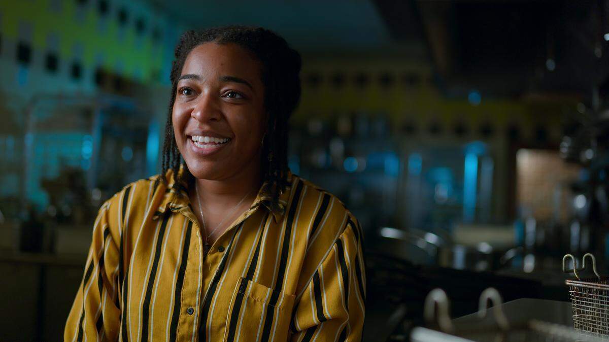 A warm, interview-style shot of a smiling young Black person with hair in braids, is wearing a yellow, black, and white vertically striped shirt. The chef is positioned in a professional kitchen, which is blurred in the background and illuminated with cool blue and green cinematic lighting.