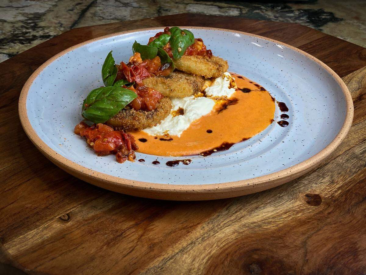 A beautifully plated Fried Green Tomato Caprese sits on a wooden board. On the speckled blue plate, a swirl of orange tomato sauce and a dollop of creamy white burrata form the base for several golden-fried green tomatoes. The stack is topped with a chunky tomato relish, large fresh basil leaves, and a drizzle of dark balsamic.