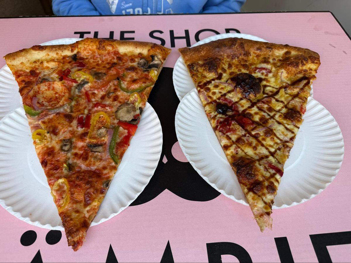 A top-down, close-up shot of two large New York-style pizza slices sitting side-by-side on white paper plates. The slice on the left is a veggie pizza loaded with melted mozzarella, tomato sauce, mushrooms, green peppers, red onions, and yellow banana peppers. The slice on the right features a golden-brown cheese base with red onions, drizzled with a crisscross pattern of dark barbecue sauce. The plates rest on a pink tabletop featuring a black graphic logo for “THE SHOP ZÄMBIES.”