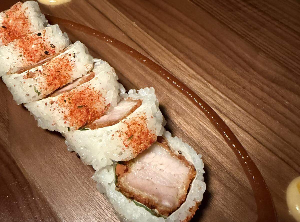 A row of six sushi rolls is arranged diagonally across a dark wood-grained surface, showcasing a fusion style of plating. Each piece features a thick, rectangular slice of breaded and fried pork belly (tonkatsu style), showing distinct layers of lean meat and fat. The filling is tightly wrapped in seasoned white sushi rice, which is seasoned with a few scattered black sesame seeds. The top of the rice is generously dusted with a bright orange-red spice blend, likely togarashi (Japanese chili flakes) or a smoky paprika.