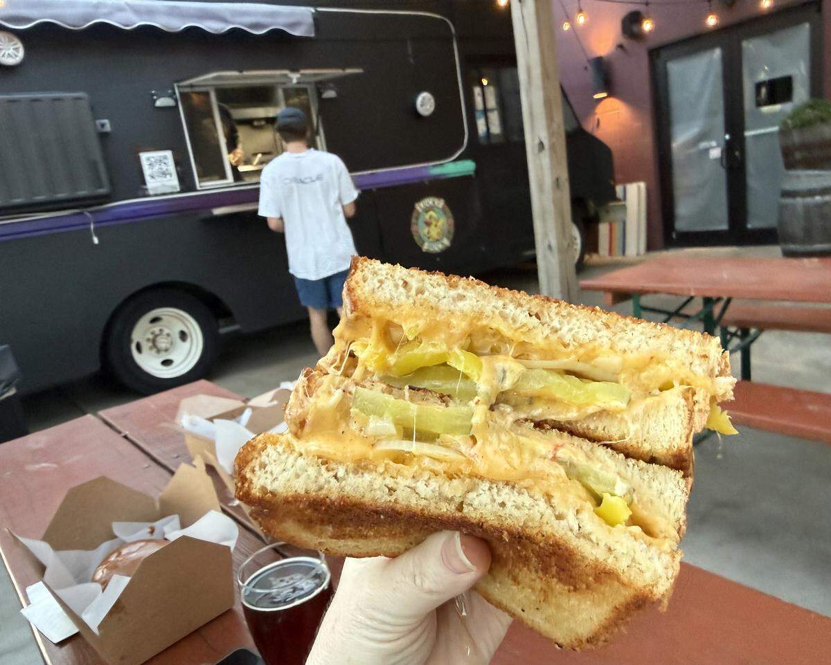 A hand holds up a thick, halved sandwich overflowing with melted orange cheese and sliced green pickles. In the blurred background, a customer stands at the serving window of a black food truck with a “Lucky Duck” logo, parked next to red picnic tables under string lights. A cardboard takeout box and a glass of dark beer sit on the table in the foreground.