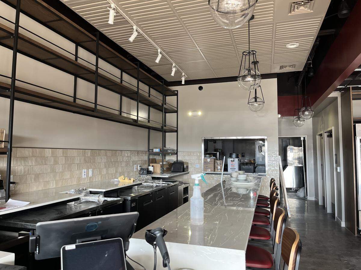A wider view of a newly built or renovated bar area. The long white counter stretches from the foreground, leading toward an open pass-through window to the kitchen area in the back. Several dark red bar stools are lined up along the counter. Above, multiple industrial-style pendant lights with clear glass globes hang from a textured white ceiling. Open metal and wood shelving lines the back wall above a subway tile backsplash.