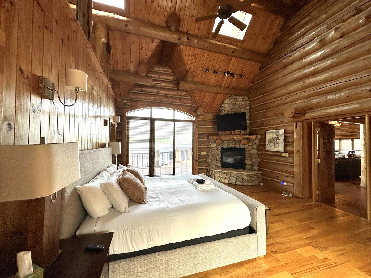 A wide view of a luxurious log cabin bedroom. The room has log walls and a high, vaulted ceiling with a skylight. In the foreground is a large, grey upholstered king bed with white linens. The bed faces a corner stone fireplace with a TV mounted above it, and beyond that, glass doors lead to a balcony overlooking a calm lake.