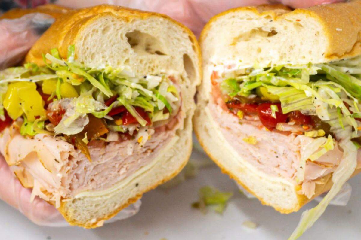 A close-up, head-on shot of a hoagie sandwich cut in half. The two halves are held up to the camera, showing the fillings of thinly sliced deli meat, cheese, shredded lettuce, and a mix of red and yellow peppers.
