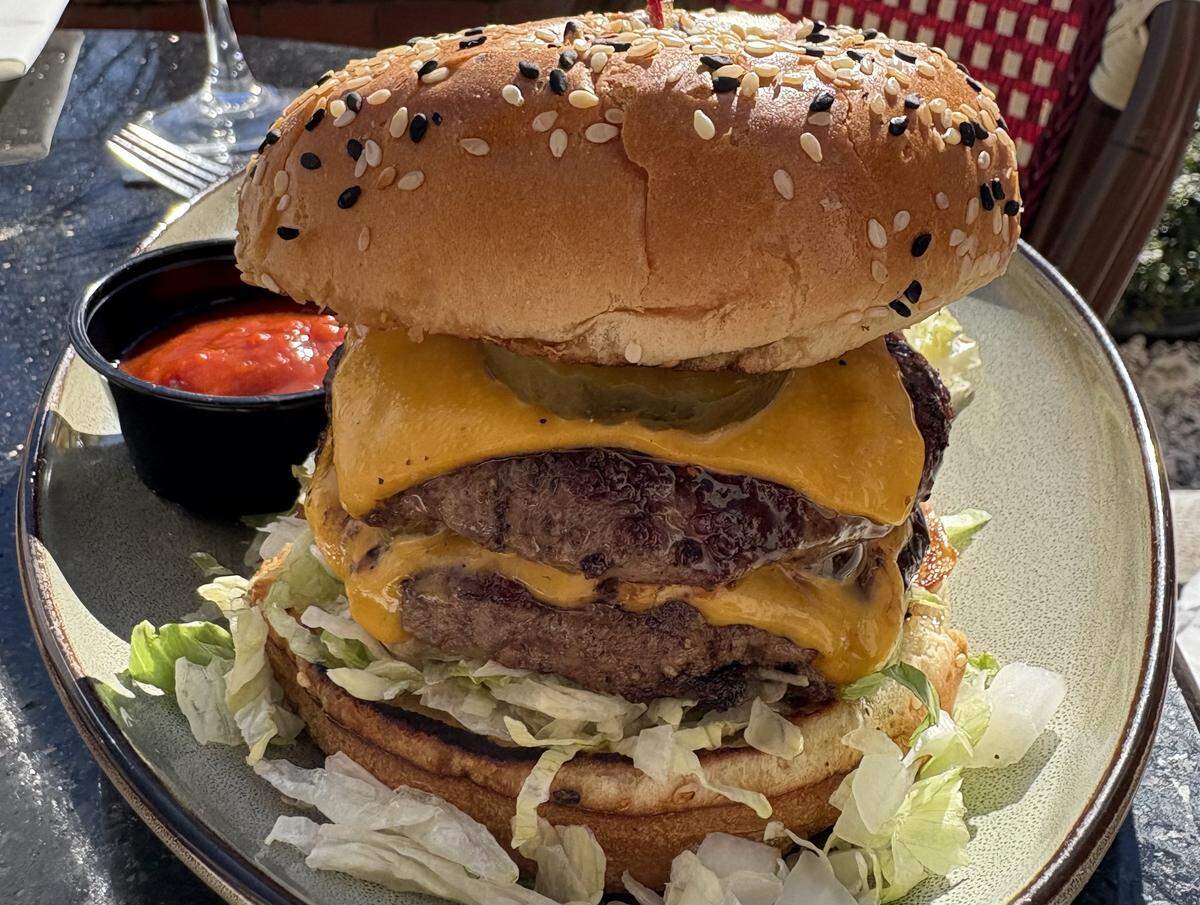 A close-up, mouth-watering shot of a double cheeseburger served on a ceramic plate. The burger features two thick, juicy beef patties layered with melted cheddar cheese, topped with a round pickle slice and shredded lettuce, all held together by a toasted everything-seasoned bun. A small black ramekin of bright red sauce or ketchup sits on the side, and the background hints at an outdoor patio setting with sunlight dappling the table.