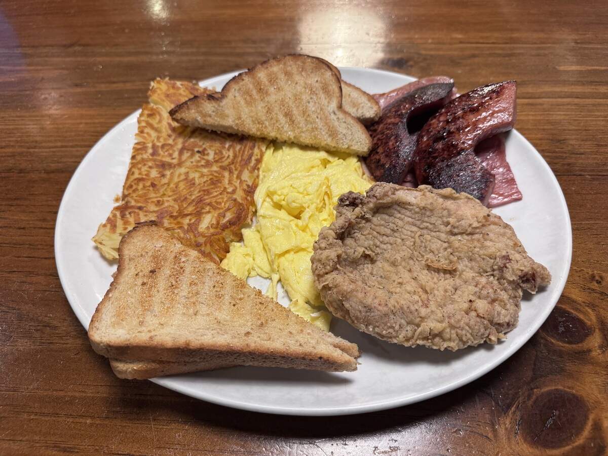 A hearty breakfast plate served on a wooden table featuring scrambled eggs, a golden-brown hash brown patty, two slices of toasted rye bread, grilled sausage slices, and a country-fried steak patty.