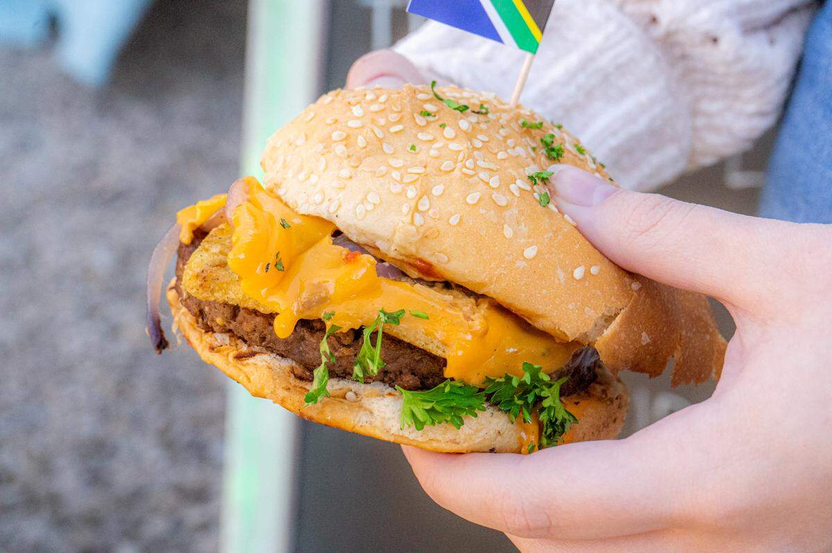 Two hands hold a single, messy vegan cheeseburger with a sesame seed bun, captured in a close-up outdoor shot. The burger features a brown patty, melted orange cheese, red onion, greens, and a small South African flag tucked into the bun.