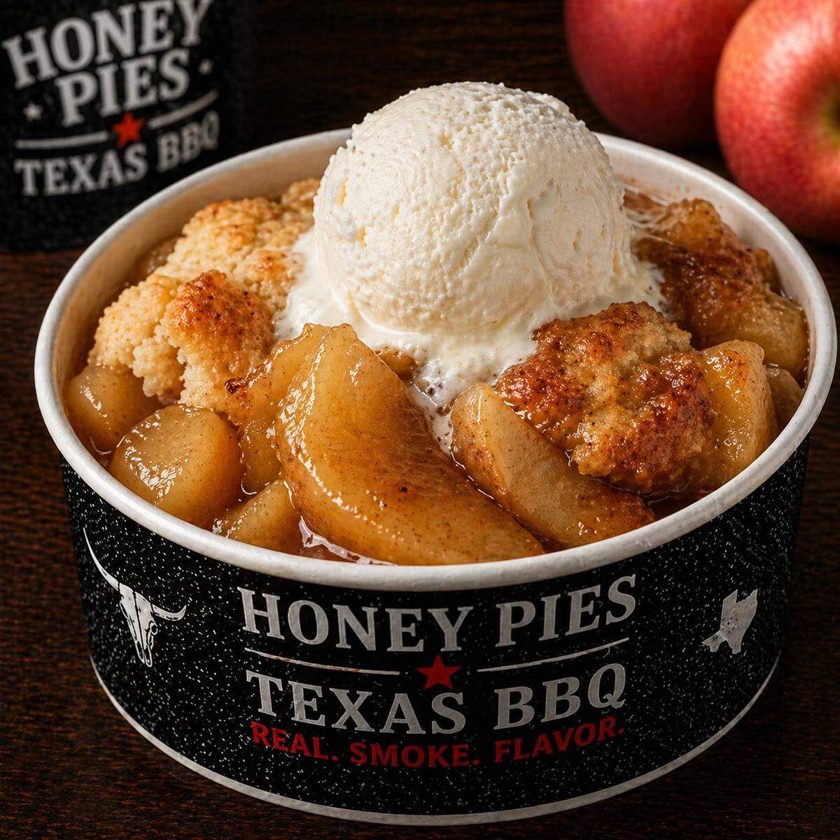 A close-up of a warm apple cobbler served in a black paper bowl with the “Honey Pies Texas BBQ” logo. The dessert features large, cinnamon-glazed apple slices and a golden biscuit topping, finished with a large scoop of melting vanilla ice cream. Red apples are visible in the blurred background.