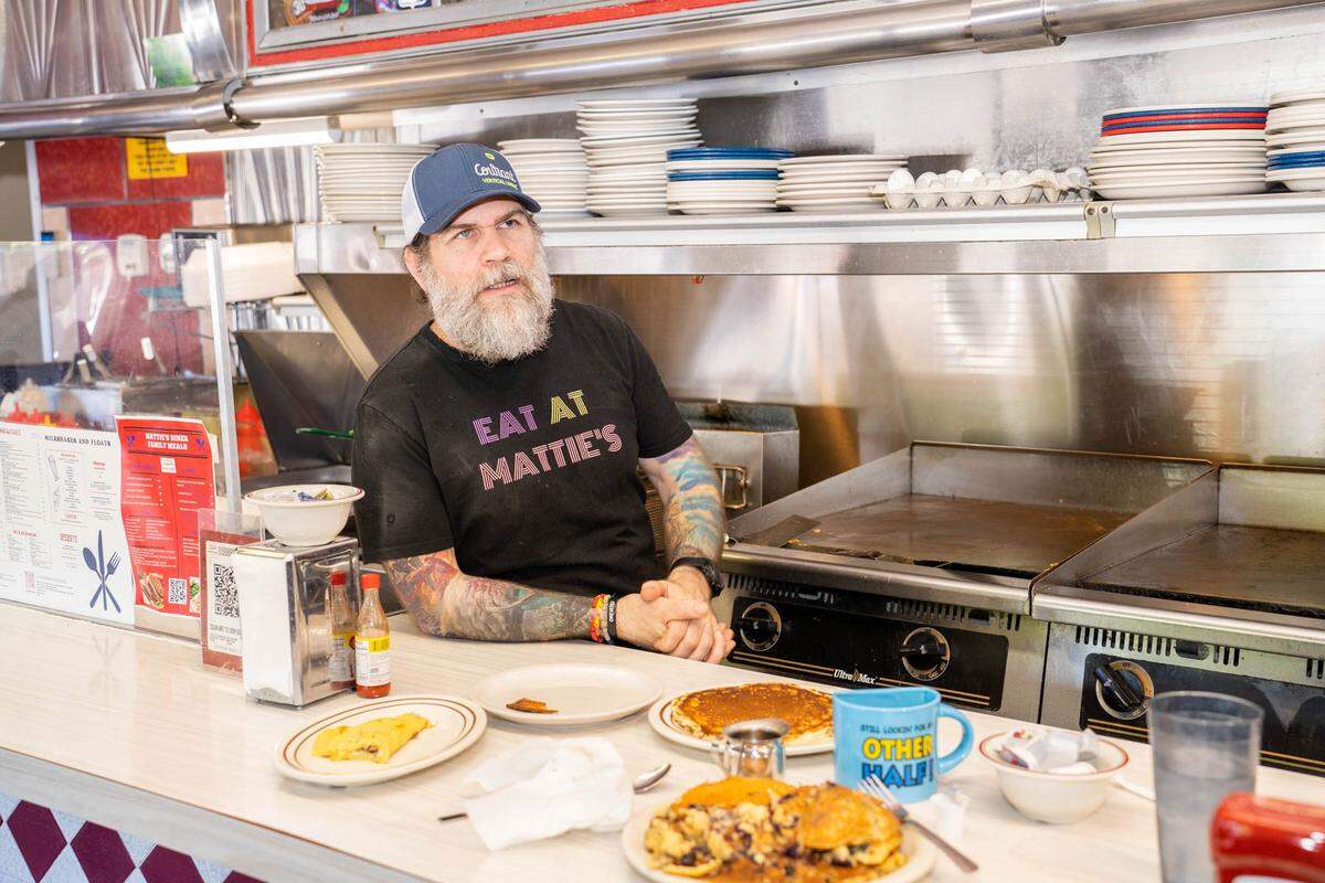 A man with a gray beard and colorful tattoos, wearing an “EAT AT MATTIE’S” t-shirt, stands behind a diner counter filled with plates of food. He is looking upward as if speaking to someone off-camera.