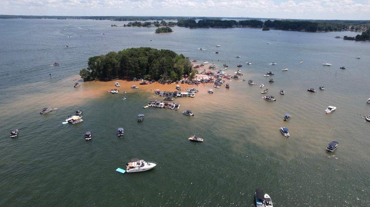 An aerial view of a large gathering of boats around a tree-covered island on a lake. Dozens of pontoon and motorboats are anchored in the shallow, sandy-colored water near the shore. The deeper water surrounding the island is a dark blue-green, and other distant islands and shorelines are visible under a bright, partly cloudy sky.